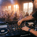 An intimate close-up shot of a DJ’s hands (face totally obscured) actively working on a glowing 4-deck Pioneer setup in a hazy club booth. It is sunrise, with golden light mixing with cool club haze through the background windows. All four vertical channel faders on the mixer are completely raised. The DJ wears headphones around their neck and is sweat-soaked, capturing the fatigue and focus of a 9-hour set.